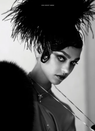 Monochromatic portrait of a woman with dark hair and dramatic eye makeup, wearing a feathered headpiece and a statement necklace.