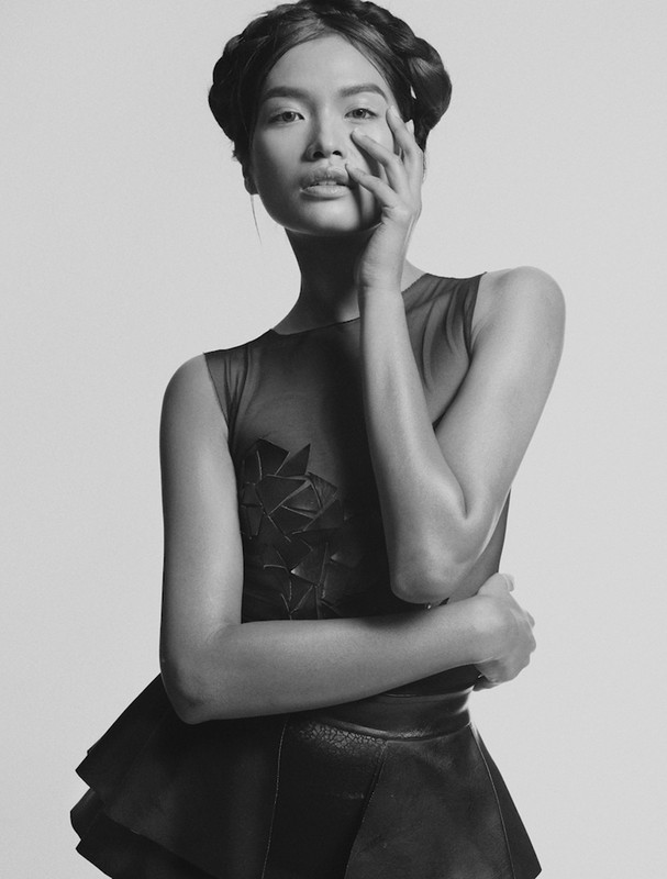 Monochrome portrait of a stylish Asian woman with hand near face, braided hair, and a geometric dress in a studio setting.