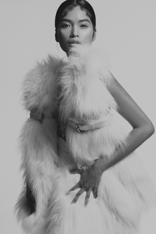 Monochrome portrait of an Asian woman in a white faux fur vest and dress, posing indoors against a neutral backdrop with serious expression.