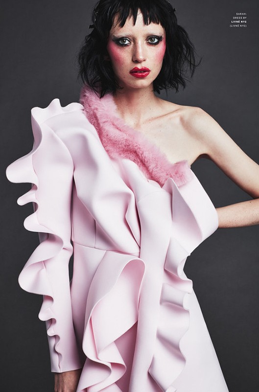 Elegant woman in a light pink, ruffled, one-shoulder dress poses against a grey background. Fashion-forward makeup and hair complete the look.