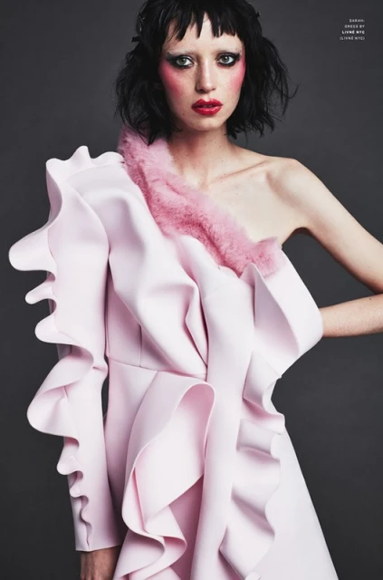 Elegant woman in a light pink, ruffled, one-shoulder dress poses against a grey background. Fashion-forward makeup and hair complete the look.