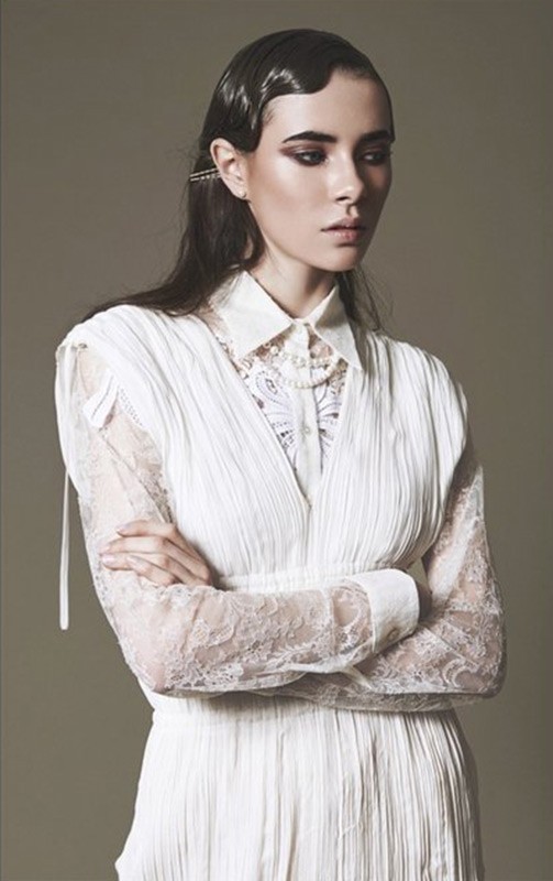 A woman poses in a white dress with a lace top and long sleeves, her arms crossed, styled hair, and dark eye makeup.