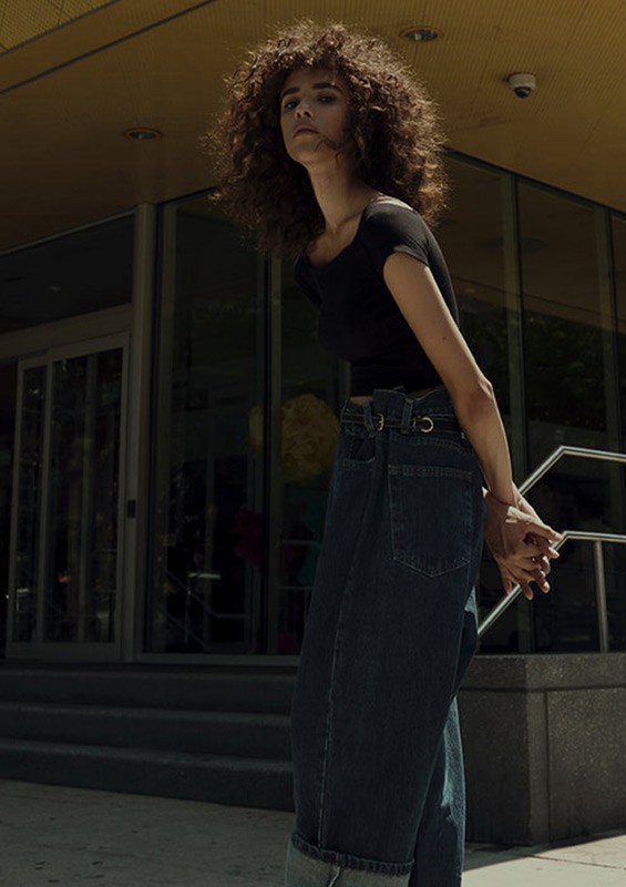 A young woman with curly hair stands in front of a building wearing a black top and wide-leg jeans on a sunny day in an urban setting.