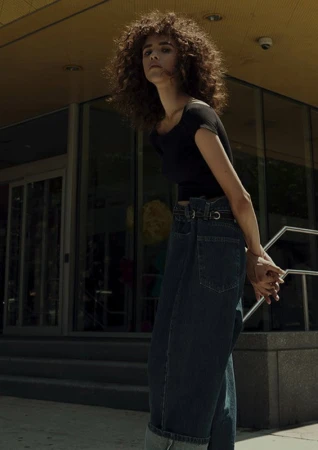 A young woman with curly hair stands in front of a building wearing a black top and wide-leg jeans on a sunny day in an urban setting.