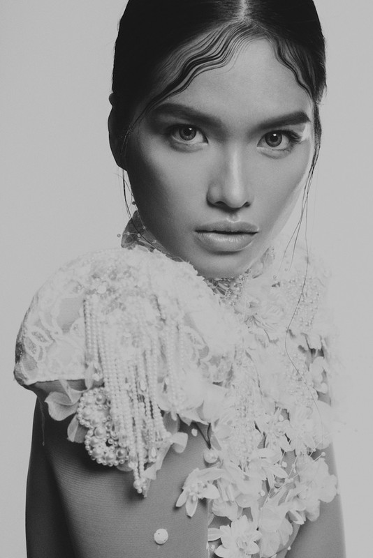 Black and white portrait of an Asian woman with art deco wavy hairstyle wearing a lace collar with pearl strands, looking at camera.