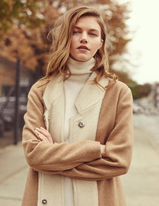 Adult female poses outdoors wearing a tan coat and cream turtleneck. The woman has long, wavy blonde hair and is facing forward.