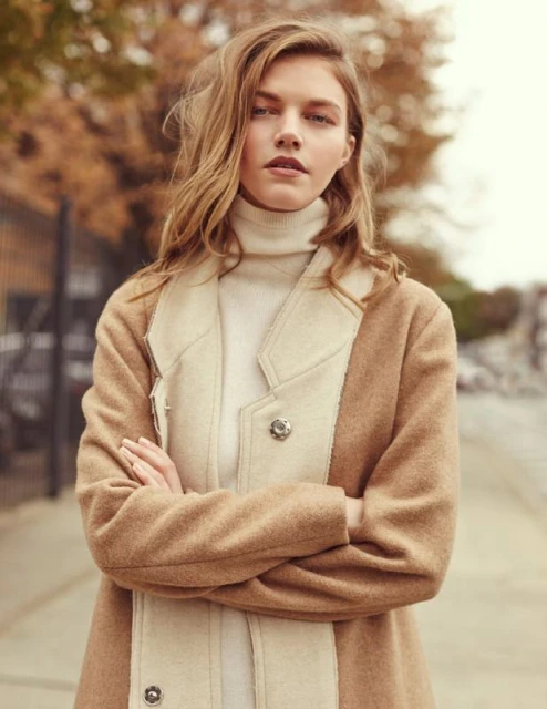 Adult female poses outdoors wearing a tan coat and cream turtleneck. The woman has long, wavy blonde hair and is facing forward.