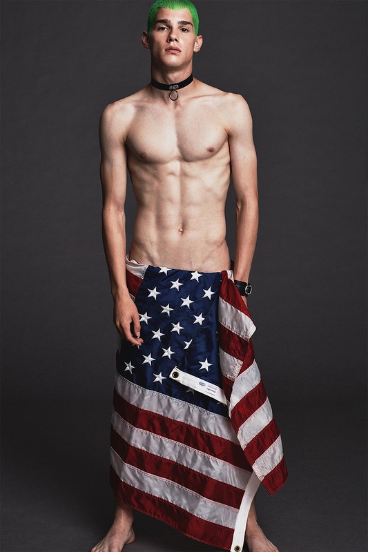 Young, shirtless man with dyed green hair stands in a photo studio partially wrapped in an American flag, accessorized with a black choker.