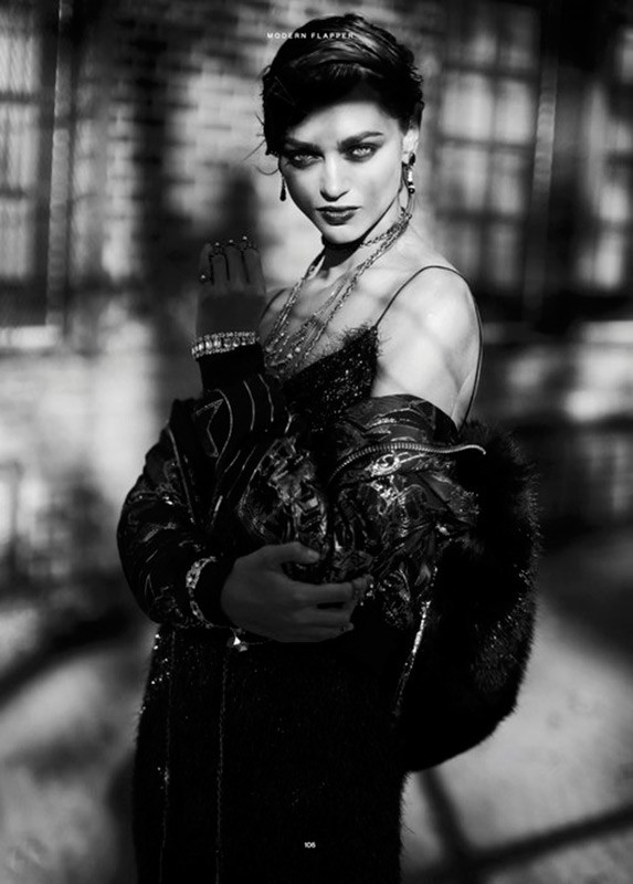 Black and white portrait of a woman wearing jewelry and a fur-lined coat, posing in front of a mottled wall.