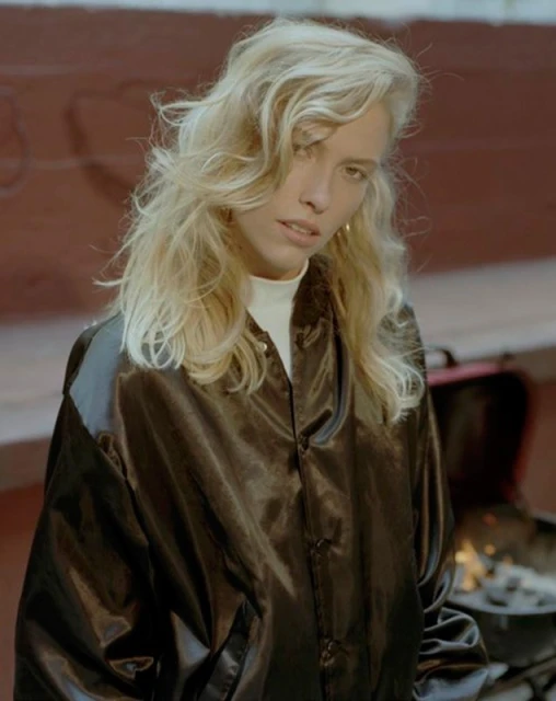 Woman with blonde wavy hair wearing a dark jacket over a white turtleneck poses outside in front of a brick wall and charcoal grill.