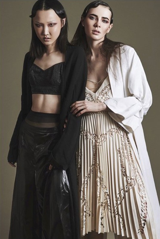Two women pose in a studio wearing black and white outfits; a dark-haired Asian woman in black and a light-skinned woman in white.