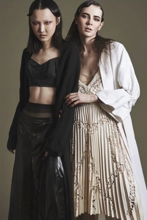 Two women pose in a studio wearing black and white outfits; a dark-haired Asian woman in black and a light-skinned woman in white.