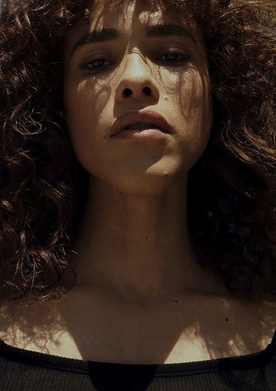 Close-up portrait of a woman with curly brown hair and dappled sunlight on her face, shot from below.