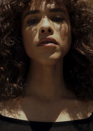 Close-up portrait of a woman with curly brown hair and dappled sunlight on her face, shot from below.