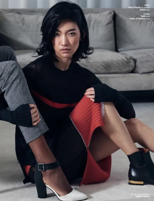 Fashion editorial shot of an Asian woman wearing a black top and red skirt, sitting with another person on a grey couch.