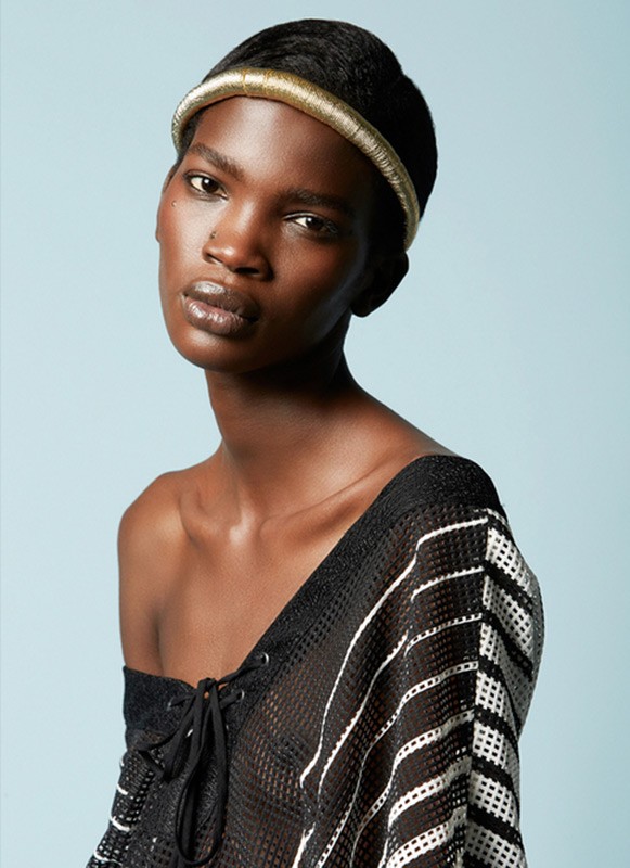 Close-up fashion portrait of a dark-skinned adult woman with a gold headband, wearing a black mesh top against a blue backdrop.
