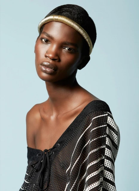 Close-up fashion portrait of a dark-skinned adult woman with a gold headband, wearing a black mesh top against a blue backdrop.