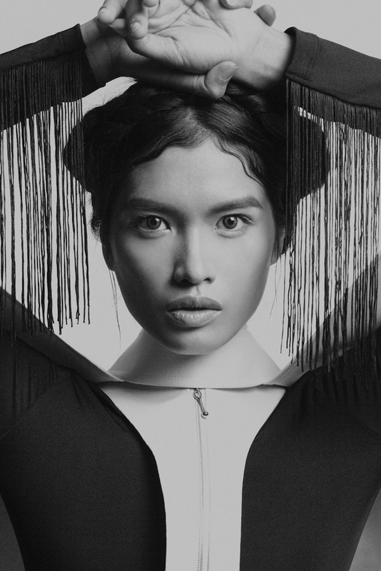 Headshot of a young woman with hands near her head, wearing a black top with fringe, shot in black and white against a light background.