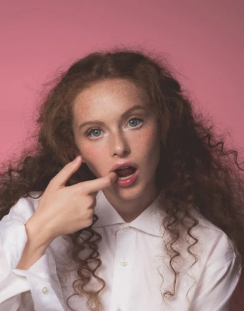 Portrait of a woman with curly brown hair, blue eyes, and freckles wearing a white buttoned shirt against a pink background.