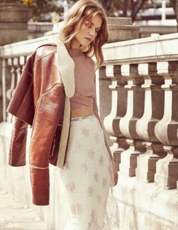 Model in mauve crop top wearing a brown leather jacket and patterned white skirt standing against an outdoor stone balustrade