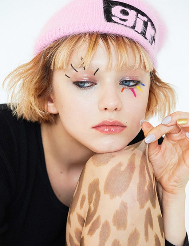 Close-up of a young woman with blonde hair and creative makeup wearing a pink beanie, resting her face on an arm with leopard print.