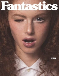Close-up portrait of a woman with curly red hair, freckles, and a winking expression wearing a white collared shirt.