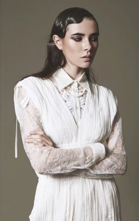 Model with slicked-back finger waves wearing a white pleated blouse with lace sleeves against a neutral grey background.