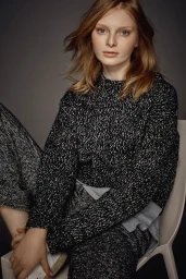 Portrait of a young woman with reddish-brown hair wearing a textured black and white sweater seated on a white chair against a dark grey background.