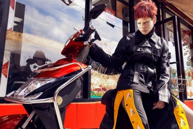 Person with red hair and bleached eyebrows posing on a red moped wearing a black leather jacket and yellow-striped pants.