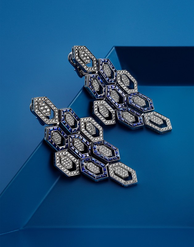 Two silver-tone statement earrings with blue and white gems, arranged in a honeycomb pattern, are displayed on a blue background.