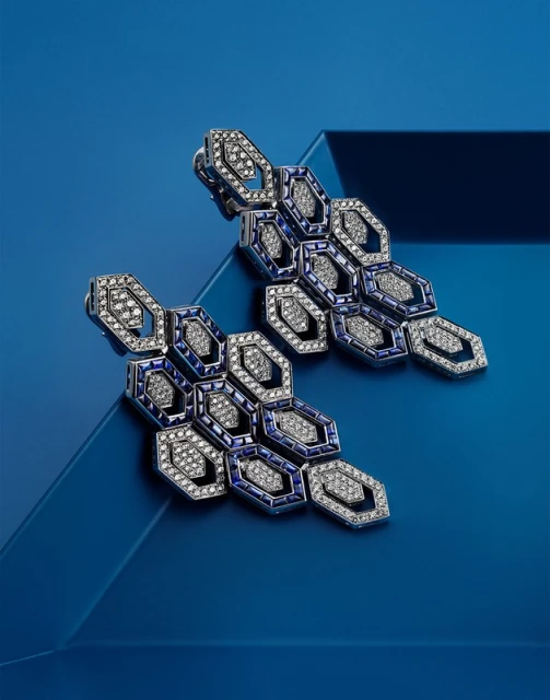 Two silver-tone statement earrings with blue and white gems, arranged in a honeycomb pattern, are displayed on a blue background.