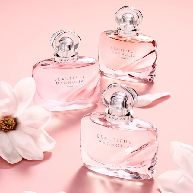 Three Estee Lauder Beautiful Magnolia perfume bottles in graduated sizes on a pink backdrop, accented with scattered white magnolia petals.