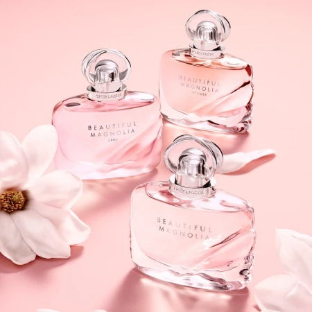 Three Estee Lauder Beautiful Magnolia perfume bottles in graduated sizes on a pink backdrop, accented with scattered white magnolia petals.