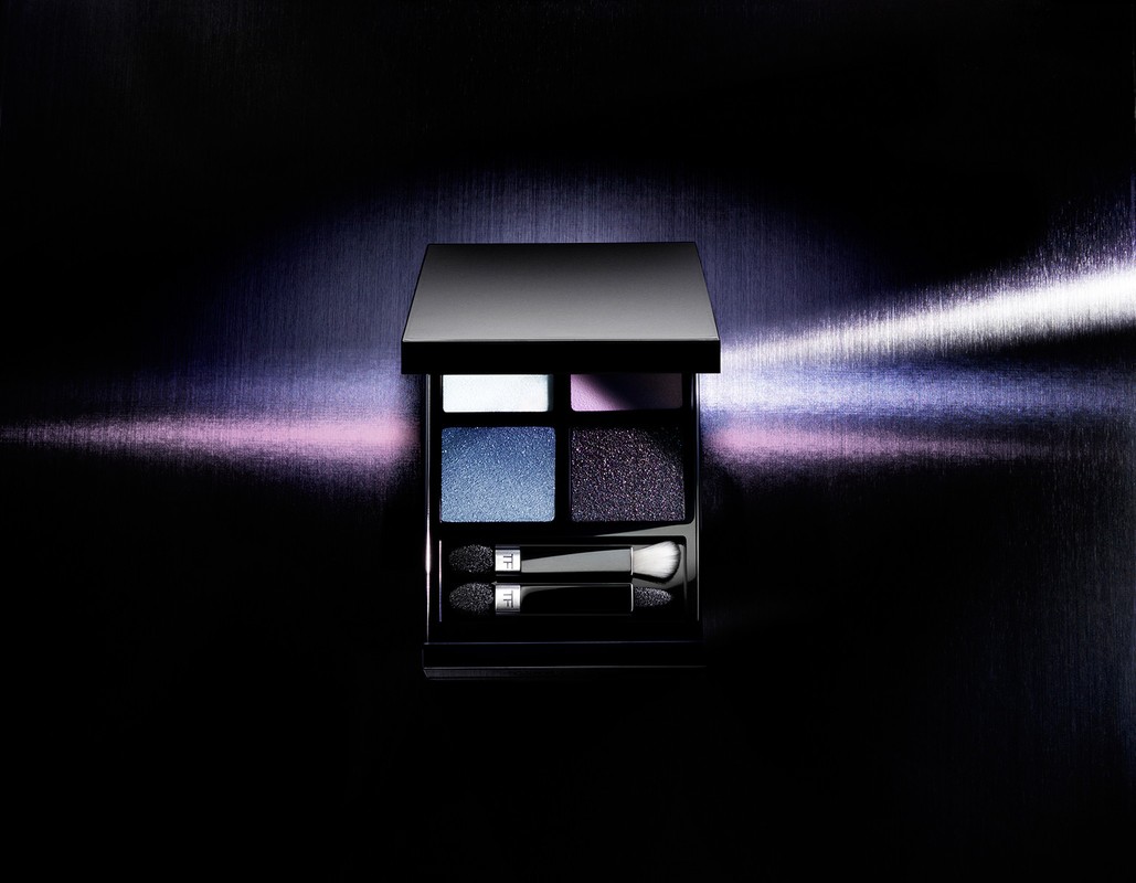 Close-up of a black eyeshadow palette with various blue and purple shades, complete with applicators, set against a dark backdrop.