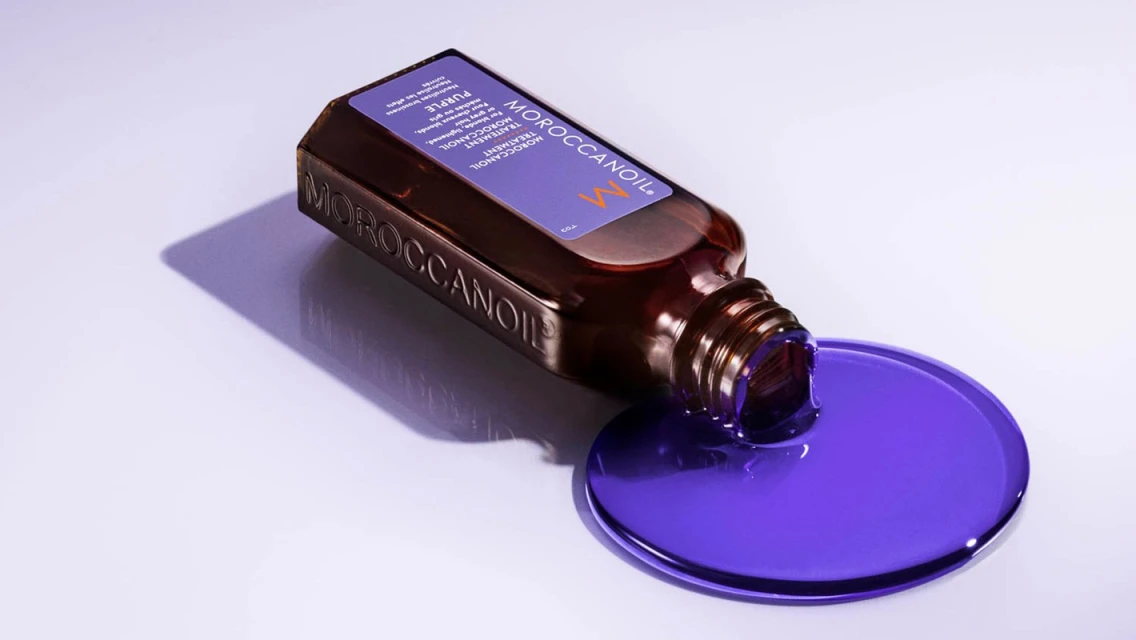 Moroccanoil Purple Treatment product lies on its side with purple liquid spilling onto a white surface in a studio setting.
