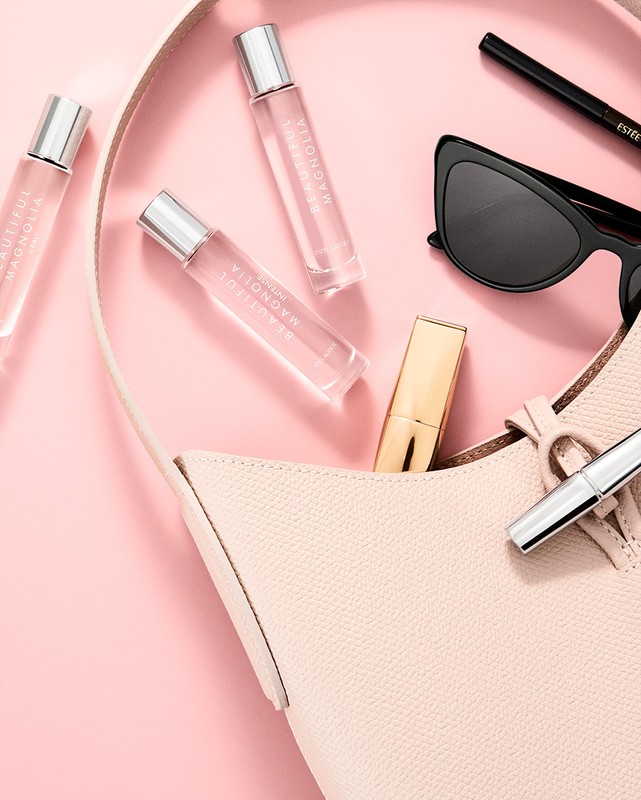 Overhead shot of a handbag with cosmetic products including perfume bottles, lipstick and sunglasses on a pink surface. Beauty editorial.