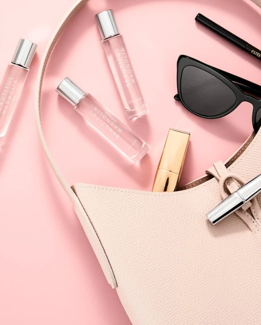 Overhead shot of a handbag with cosmetic products including perfume bottles, lipstick and sunglasses on a pink surface. Beauty editorial.
