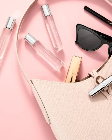 Overhead shot of a handbag with cosmetic products including perfume bottles, lipstick and sunglasses on a pink surface. Beauty editorial.