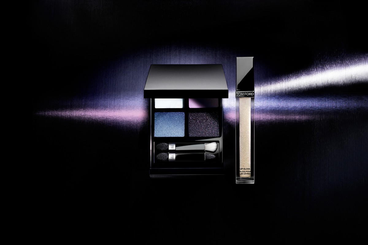 Tom Ford eyeshadow quad and lip gloss displayed against a dark metallic background with horizontal light beam.
