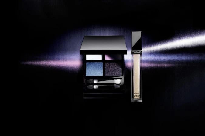Tom Ford eyeshadow quad and lip gloss displayed against a dark metallic background with horizontal light beam.