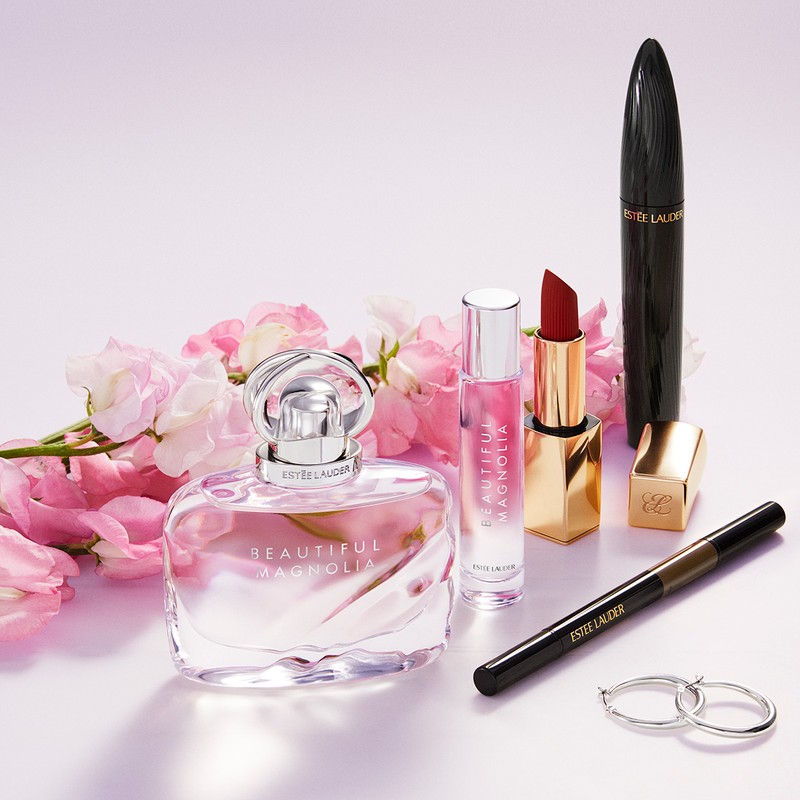 Estee Lauder beauty product arrangement: Beautiful Magnolia perfume, lipstick, mascara, eyeliner, earrings, and pink floral accents.