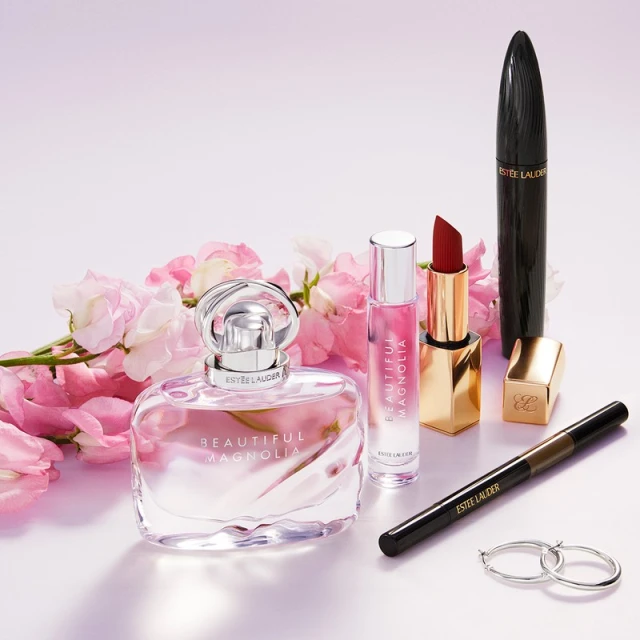 Estee Lauder beauty product arrangement: Beautiful Magnolia perfume, lipstick, mascara, eyeliner, earrings, and pink floral accents.