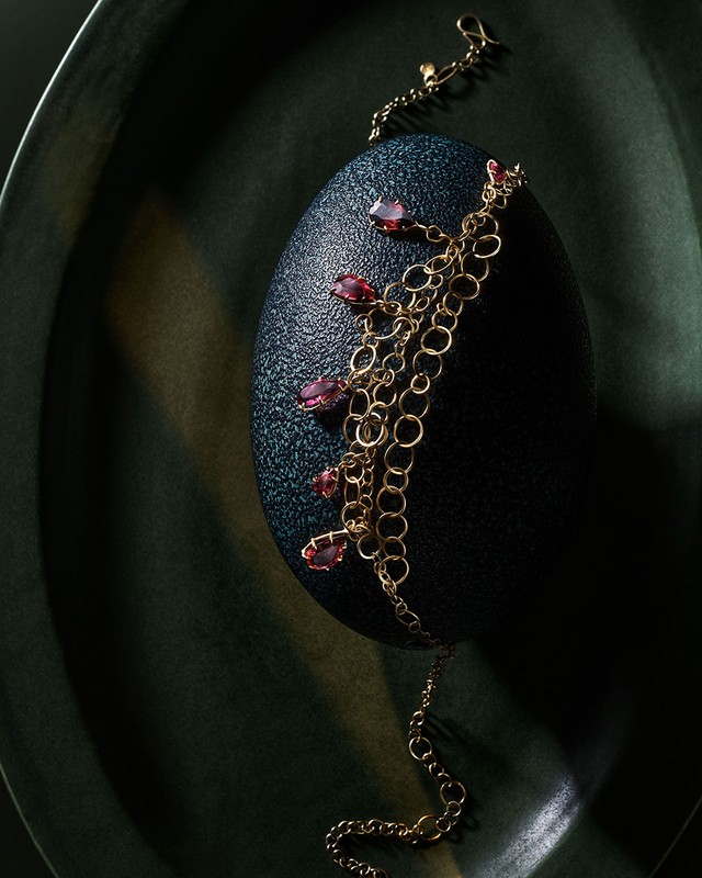 Elegant gold chain bracelet with red gemstones displayed on dark textured egg-shaped surface, presented on a dark green textured plate.
