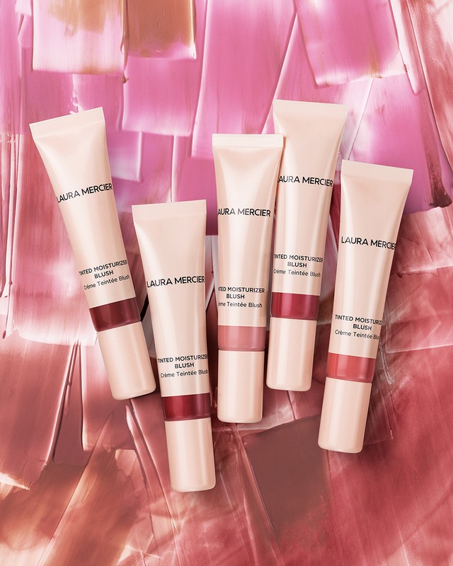 Laura Mercier tinted moisturiser blush tubes are arranged on a textured pink surface in a beauty product shot, showcasing cosmetic packaging.