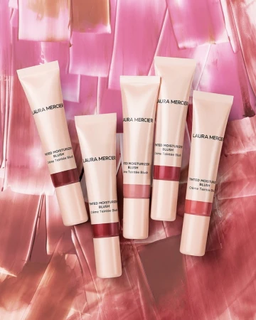 Laura Mercier tinted moisturiser blush tubes are arranged on a textured pink surface in a beauty product shot, showcasing cosmetic packaging.