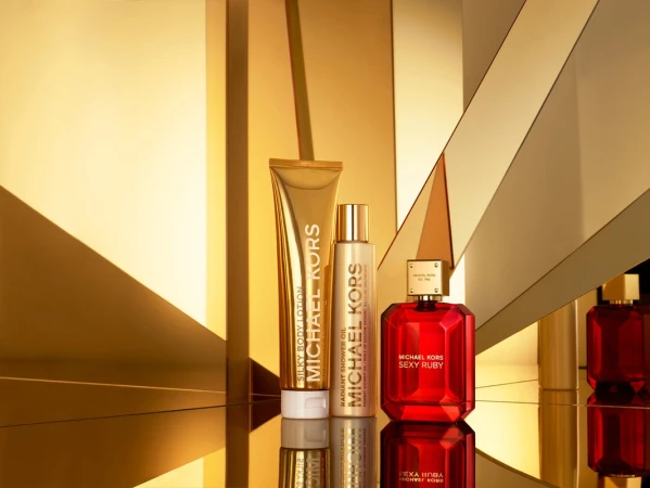 Michael Kors beauty products featuring silky body lotion, radiant shower oil, and sexy ruby perfume against a reflective gold backdrop.