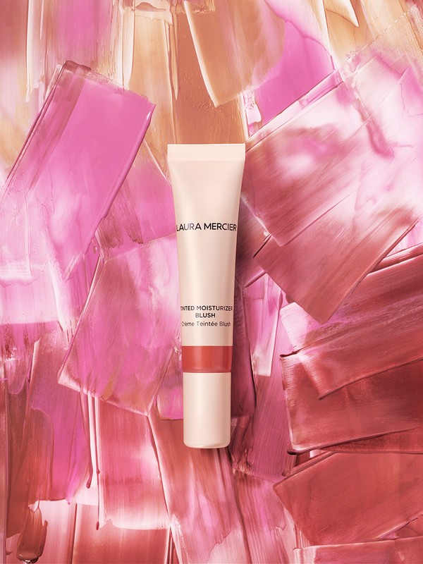 Tube of Laura Mercier Tinted Moisturizer Blush against a backdrop of pink, peach, and cream brushstroke textures. Beauty product shot.