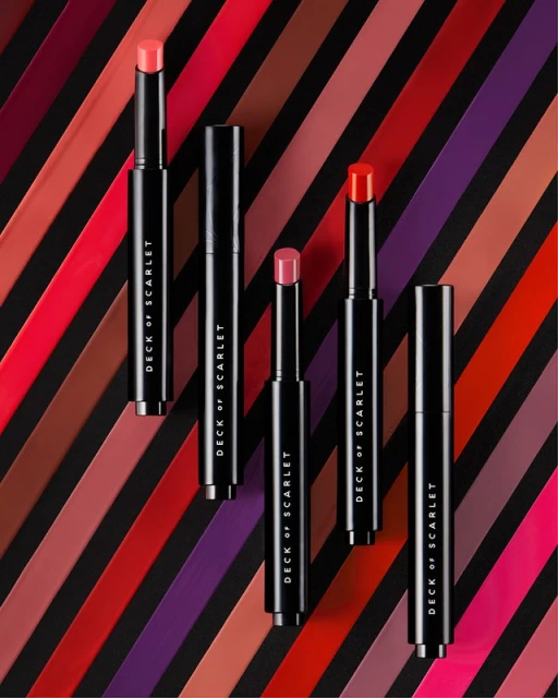 Cosmetics still life featuring several Deck of Scarlet lipstick tubes with varying color caps against a diagonally striped background.