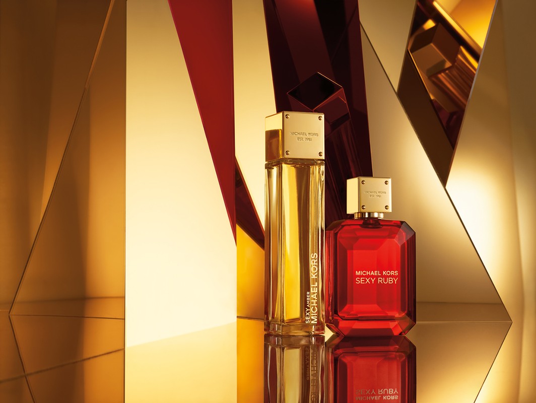 Two Michael Kors perfume bottles, Sexy Amber and Sexy Ruby, displayed on a reflective surface against a geometric gold background.