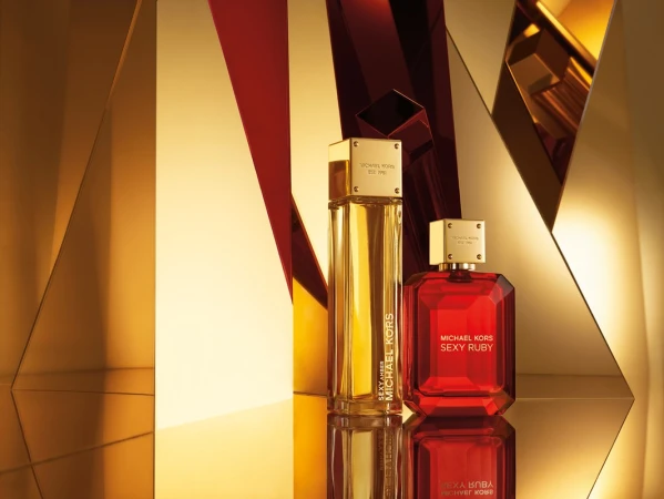 Two Michael Kors perfume bottles, Sexy Amber and Sexy Ruby, displayed on a reflective surface against a geometric gold background.